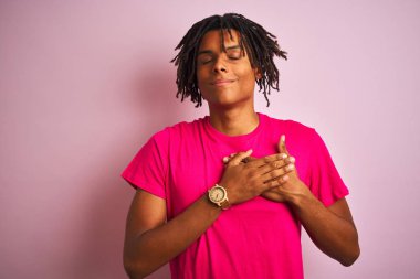 Rastalı tişörtlü Afro-Amerikalı adam izole edilmiş pembe arka planda dikiliyor elleri göğsünde, gözleri kapalı ve yüzünde minnet dolu bir jest ile gülümsüyor. Sağlık kavramı.