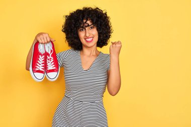 Kıvırcık saçlı genç Arap kadın izole edilmiş sarı arka planda spor ayakkabılarını tutuyor gururla bağırıyor ve zaferi ve başarıyı kutluyor.