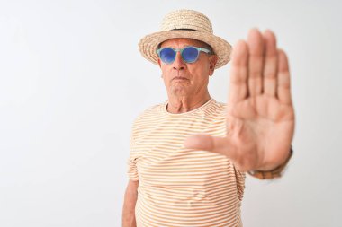 Kıdemli adam çizgili tişört gözlük takıyor, güneş gözlüğü takıyor ve izole edilmiş beyaz arka plan üzerine süvari şapkası takıyor. Açık elle dur işareti yapıyor. Ciddi ve kendinden emin bir ifadeyle, savunma jesti.