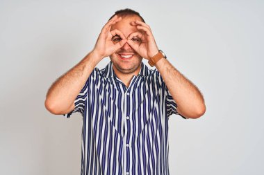 İzole edilmiş beyaz arka plan üzerinde duran mavi çizgili bir gömlek giyen genç adam dürbün gibi dil çıkartıyor, gözler parmaklarından bakıyor. Çılgın ifade.