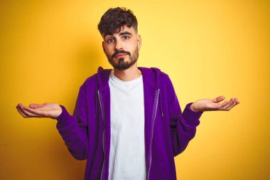 İzole edilmiş sarı arkaplan üzerine mor kazak giyen, kolları ve elleri havada, kafası karışmış, dövmeli genç bir adam. Şüphe kavramı.