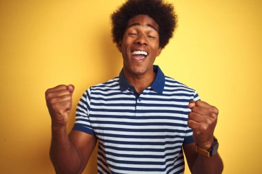 Afro saçlı, lacivert çizgili polo giyen Amerikalı bir adam izole edilmiş sarı arka planda çok mutlu ve heyecanlı bir şekilde kollarını kaldırmış, gülümseyen ve başarı için haykıran bir jest yapıyor. Kutlama konsepti.