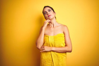 Young beautiful woman wearing towel after shower over yellow isolated background with hand on chin thinking about question, pensive expression. Smiling with thoughtful face. Doubt concept.