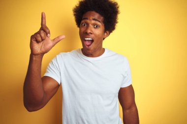 Siyah saçlı, beyaz tişörtlü, izole edilmiş sarı arka planda duran Amerikalı bir adam başarılı bir fikri işaret ediyor. Heyecanlı ve mutlu. Bir numara..