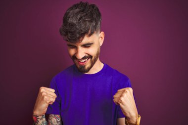 İzole edilmiş mor arka plan üzerinde duran dövmeli genç adam mutlu ve heyecanlı bir şekilde kollarını kaldırıp, gülümseyerek ve başarı için çığlık atarak zafer hareketi yapıyor. Kutlama konsepti.
