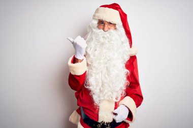 Noel Baba kostümü giyen orta yaşlı yakışıklı adam izole edilmiş beyaz arka planda gülümseyen ve baş parmağıyla yüzünü gösteren..