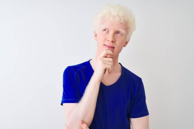Mavi günlük tişört giyen genç albino bir adam izole edilmiş beyaz arka plan üzerinde duruyor ve elleri çenesinde soru sormayı düşünüyor, düşünceli bir ifade. Düşünceli bir yüzle gülümsüyor. Şüphe kavramı.