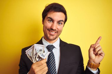 Soyutlanmış sarı arka planda dolar tutan takım elbiseli yakışıklı bir işadamı. Elleri ve parmakları kenarda mutlu mesut işaret ediyor.
