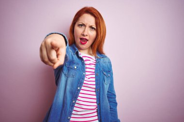 Güzel kızıl saçlı kadın, kot gömlek ve çizgili tişört giyip pembe arka planda izole edilmiş. Kameraya kızgın ve sinirli olduğunu gösteriyor.
