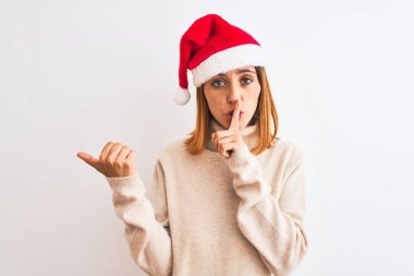 Güzel kızıl saçlı bir kadın, izole edilmiş arka planda noel şapkası takıyor ve sessiz olmak istiyor. Parmakları dudaklarında, elleri yandan işaret ederek. Sessizlik ve gizli konsept.
