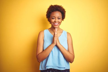Güzel Afro-Amerikalı kadın zarif bir tişört giyerek izole edilmiş sarı arka planda dua ederek ellerini birleştirip af diliyor kendinden emin bir şekilde gülümsüyor..