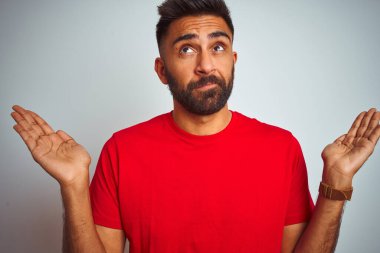 İzole edilmiş beyaz arka plan üzerine kırmızı t-shirt giyen genç Hintli adam elleri ve kolları havada, kafası karışık bir ifade takındı. Şüphe kavramı.