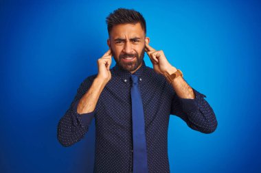 Genç Hintli iş adamı zarif gömlek ve kravat giyerek izole edilmiş mavi arka planda duruyor. Yüksek sesli müzik sesi için parmaklarıyla kulaklarını kapatıyor. Sağır kavramı.