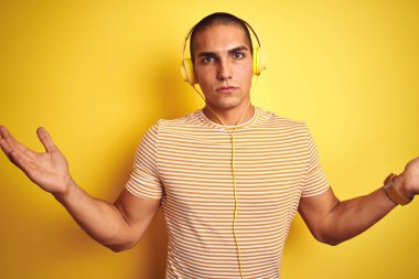 Sarı arka plan üzerinde kulaklık kullanan genç yakışıklı bir adam. Kolları ve elleri havada, kafası karışmış bir yüz ifadesi. Şüphe kavramı.