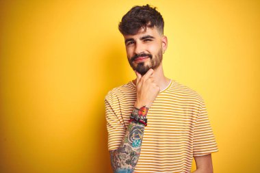 İzole edilmiş sarı arka plan üzerinde duran çizgili tişörtlü genç adam çapraz kollarla gülümseyen ve elleri çenesinde olan kameraya bakıyor. Olumlu düşünüyorum..