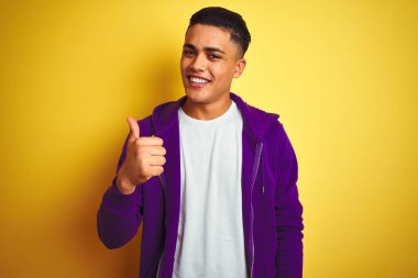 Young brazilian man wearing purple sweatshirt standing over isolated yellow background doing happy thumbs up gesture with hand. Approving expression looking at the camera showing success. clipart