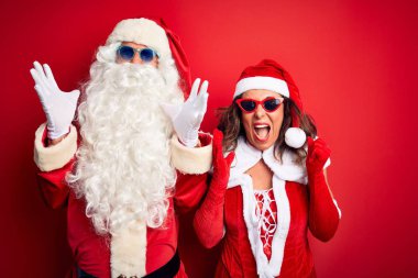 Middle age couple wearing Santa costume and sunglasses over isolated red background celebrating mad and crazy for success with arms raised and closed eyes screaming excited. Winner concept clipart