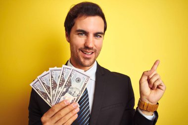 Soyutlanmış sarı arka planda dolar tutan takım elbiseli yakışıklı bir işadamı. Elleri ve parmakları kenarda mutlu mesut işaret ediyor.
