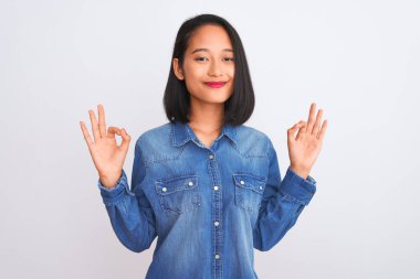 İzole edilmiş beyaz arka plan üzerinde duran kot gömlek giyen genç ve güzel Çinli kadın rahatla ve gözleri kapalı parmaklarıyla meditasyon hareketi yaparak gülümse. Yoga konsepti.