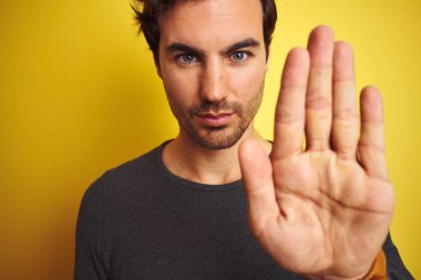 Sarı arka plan üzerinde duran, ciddi ve kendinden emin ifadesiyle dur işareti yapan, sıradan bir süveter giyen genç yakışıklı bir adam.