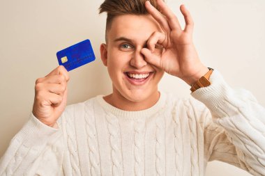Genç ve yakışıklı bir adam kredi kartını izole edilmiş beyaz arka planda tutarken gülümseyen yüzüyle tamam işareti yaparken parmaklarının arasından bakıyor.
