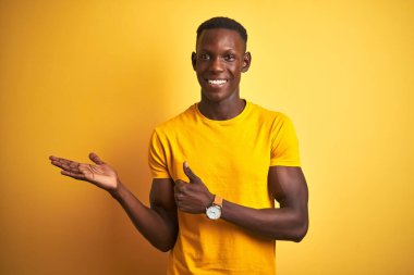 Genç Afro-Amerikalı bir adam günlük tişört giyip izole edilmiş sarı arka planda duruyor, avuç içini gösteriyor ve baş parmaklarıyla iyi bir hareket yapıyor, mutlu ve neşeli gülümsüyor.