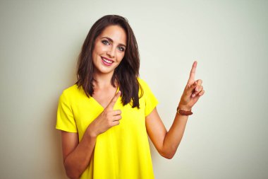 Beyaz izole arka plan üzerinde duran sarı t-shirt giyen genç güzel kadın gülümseyerek ve yan iki el ve parmakları ile işaret kamera bakıyor.