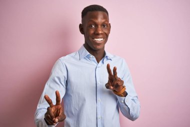 İzole edilmiş pembe arka planda duran mavi şık gömlekli Afro-Amerikalı adam kameraya bakıp zafer işareti yaparken gülümsüyor. İki numara..