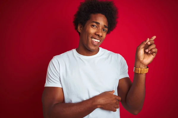 Young american man with afro hair wearing white t-shirt standing over isolated red background with a big smile on face, pointing with hand and finger to the side looking at the camera.