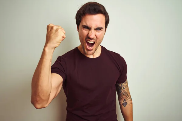 stock image Young handsome man with tattoo wearing purple casual t-shirt over isolated white background angry and mad raising fist frustrated and furious while shouting with anger. Rage and aggressive concept.
