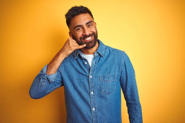 デニムのシャツを着た若いインド人男性が 電話で話すような手や指で携帯電話のジェスチャーをして笑って孤立した黄色の背景の上に立っている 概念の伝達 — ストック写真