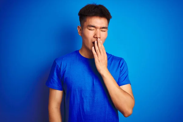 年轻的亚裔男子穿着T恤衫站在孤立的蓝色背景下 厌烦了打呵欠累了用手捂住嘴 烦躁不安和困倦 — 图库照片