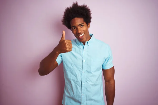 年轻的美国人 头发是非洲裔 穿着蓝色的衬衫 站在孤立的粉色背景上 手举着快乐的大拇指 在镜头前看着成功的表情 — 图库照片
