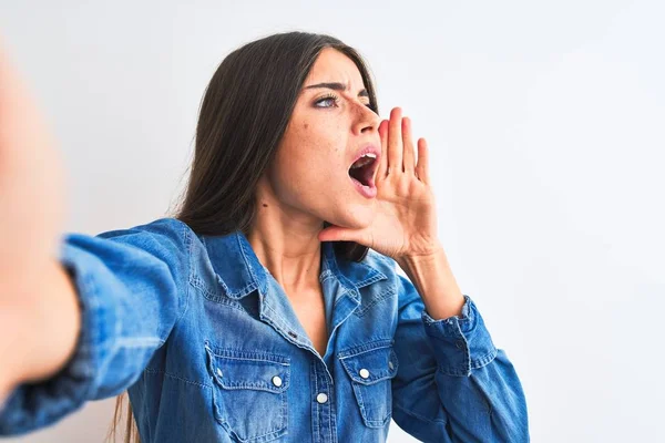 Beautiful woman wearing denim shirt make selfie by camera over isolated white background shouting and screaming loud to side with hand on mouth. Communication concept.