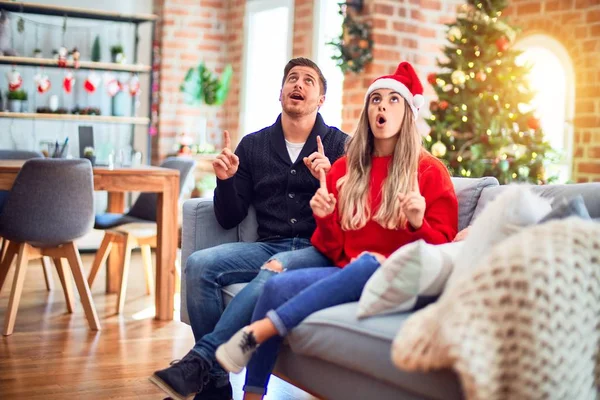 一对年轻的夫妇 头戴圣爪帽 坐在沙发上 围着圣诞树坐在家里 惊讶地抬起头 用手指和胳膊指指着 — 图库照片
