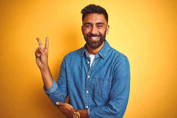 独立した黄色の背景にデニムシャツを着た若いインド人男性が 勝利サインをするカメラで幸せそうな顔をして笑っている 第二番 — ストック写真