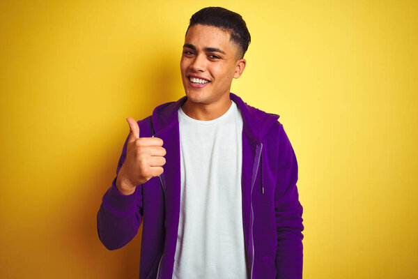 Young brazilian man wearing purple sweatshirt standing over isolated yellow background doing happy thumbs up gesture with hand. Approving expression looking at the camera showing success.