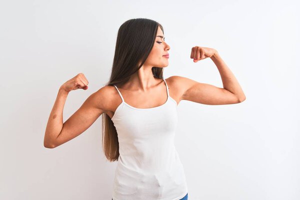 Young beautiful woman wearing casual t-shirt standing over isolated white background showing arms muscles smiling proud. Fitness concept.