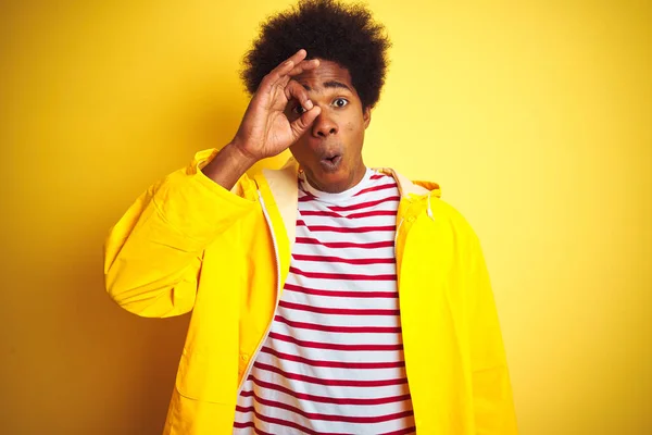 African american man with afro hair wearing rain coat standing over isolated yellow background doing ok gesture shocked with surprised face, eye looking through fingers. Unbelieving expression.