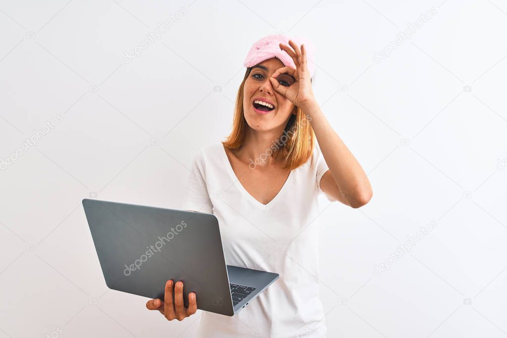 Beautiful redhead woman wearing sleeping mask using laptop over isolated background with happy face smiling doing ok sign with hand on eye looking through fingers