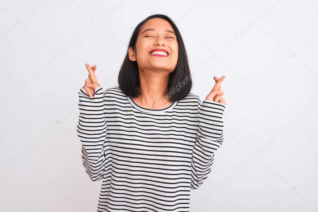 Young chinese woman wearing striped t-shirt standing over isolated white background gesturing finger crossed smiling with hope and eyes closed. Luck and superstitious concept.