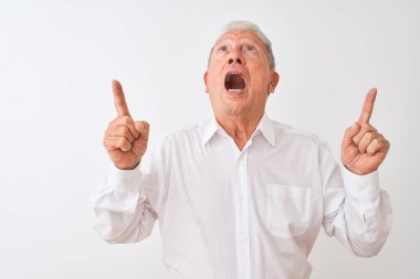 Üst düzey gri saçlı, zarif tişörtlü adam izole edilmiş beyaz arka planda duruyor. Şaşırmış ve şaşırmış bir şekilde yukarı bakıyor ve parmaklarıyla işaret ediyor ve kollarını kaldırıyor..