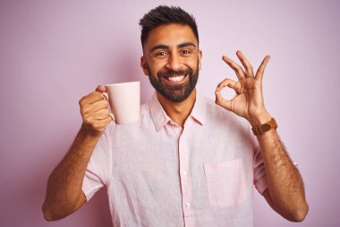 Genç Hintli adam gömleğini giyiyor kahve içiyor pembe arka planda duruyor tamam işareti yapıyor, mükemmel bir sembol.