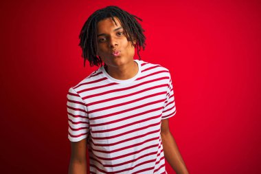 Rastalı tişörtlü genç afro bir adam izole edilmiş kırmızı arka planda dikilip kameraya bakıyor ve güzel ve seksi bir şekilde öpüşüyor. Aşk ifadesi.