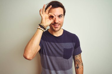 İzole edilmiş beyaz arka plan üzerine gündelik tişört giyen genç yakışıklı adam el hareketleriyle gülümsüyor ve mutlu yüzle parmaklarının arasından bakıyor..