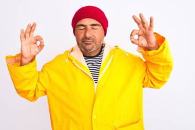 Yağmurluk ve yünlü şapka giyen orta yaşlı bir adam izole edilmiş beyaz arka planda dikiliyor rahatlıyor ve parmaklarıyla meditasyon hareketi yaparken gözleri kapalı gülümsüyor. Yoga konsepti.