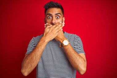 İzole edilmiş kırmızı arka plan üzerinde denizci çizgili tişört giyen genç ve yakışıklı Hintli bir adam ağzını yanlışlıkla ellerle kapatarak şok etti. Gizli konsept.
