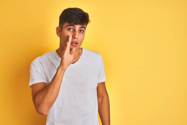 Young indian man wearing white t-shirt standing over isolated yellow background hand on mouth telling secret rumor, whispering malicious talk conversation
