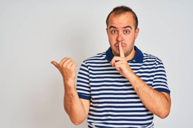 Sıradan çizgili polo giyen genç bir adam izole edilmiş beyaz arka planda dikiliyor ve parmakları yandan kenarı işaret eden dudaklarıyla sessiz olmak istiyor. Sessizlik ve gizli konsept.