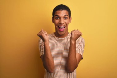 İzole edilmiş sarı arka plan üzerinde duran çizgili tişört giyen genç yakışıklı Arap adam kollarını kaldırarak ve açık gözlerle başarının şaşkınlığını ve şaşkınlığını yaşıyor. Kazanan konsept.